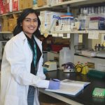 Portrait of Khushali Trivedi in a lab. She is wearing a lab coat. She is holding a pen and a note book is on the counter.