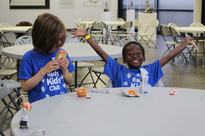 One child smiles with his arms stretched out. Another child drinks from a juice box.