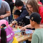 A university students with long hair and a red shirt working with colorful plastic straws to build structures and cubes while explaining it to the kids that are gathered around the table.