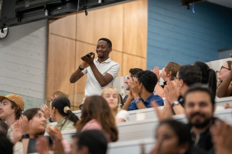 A student standing at an amphitheatre style classroom, surrounded by other students who are smiling and clapping.